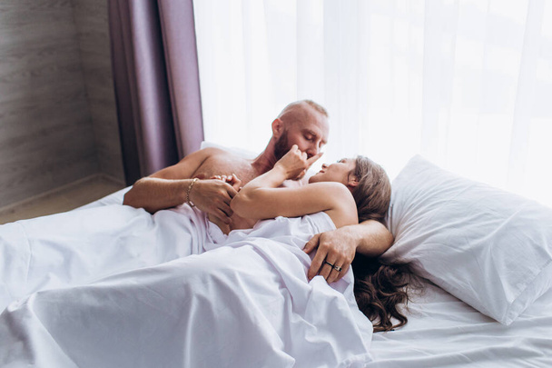 愛情のあるカップルがベッドで笑う。白いシーツの上で笑顔. - 写真・画像
