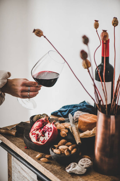 Red wine, board with cheese, fruit, almonds and olives, decorative flowers on kitchen counter and womans hand with glass of wine, white wall background. Wine tasting, wine bar concept - Foto, Imagem