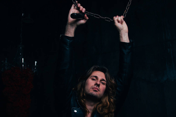brutal man in a leather jacket on a naked body with long hair. a lonely man is sad and desperate. holds a microphone in his hand took up the steel chain, - Valokuva, kuva