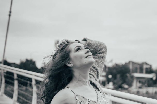 όμορφο κορίτσι στέκεται στη γέφυρα, ο αέρας φυσά στο πρόσωπό της, αναπτύσσοντας τα μαλλιά της. κορίτσι χαμόγελα. χορός ασπρόμαυρη φωτογραφία. - Φωτογραφία, εικόνα