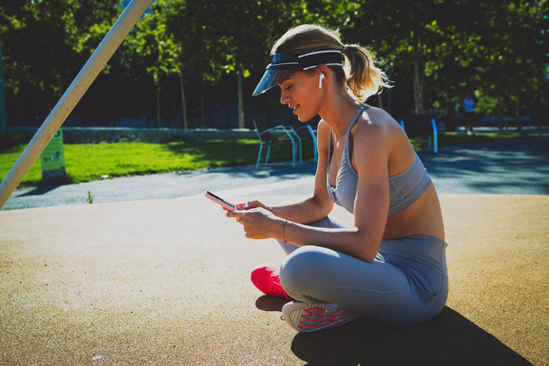 Side view portrait of the athletic young female with earphones using mobile phone while sitting on a sports ground with her legs crossed. Sporty woman checking training results in a mobile phone app - Fotografie, Obrázek