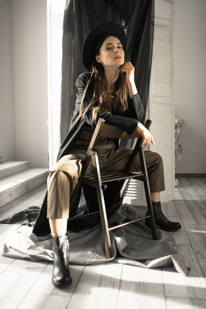 attraktive brünette Frau in modischer Kleidung sitzt auf Stuhl und posiert im Studio  - Foto, Bild