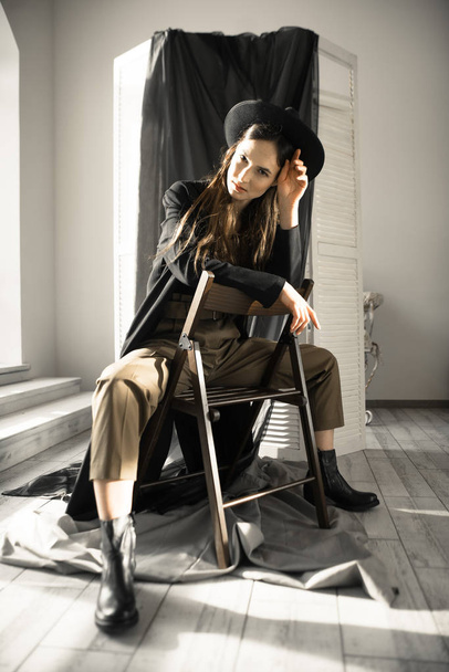 atractiva mujer morena en ropa de estilo de moda sentado en la silla y posando en el estudio
  - Foto, Imagen