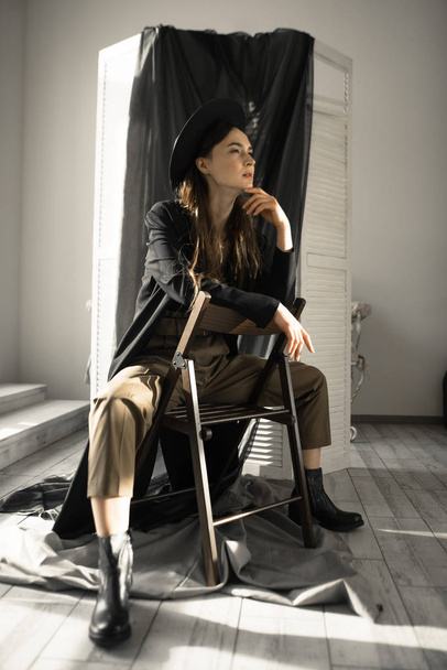 attraktive brünette Frau in modischer Kleidung sitzt auf Stuhl und posiert im Studio  - Foto, Bild