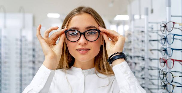 Happy female client or optician is standing with set of glasses in background in optical shop. Woman in spectacles. Girl wearing glasses. Portrait of a woman in correcting glasses. Closeup. - Valokuva, kuva