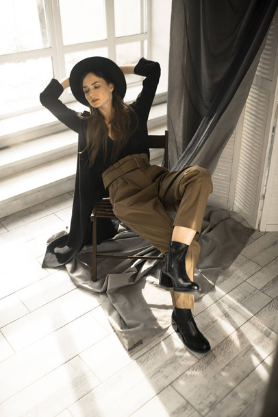 atractiva mujer morena en ropa de estilo de moda sentado en la silla y posando en el estudio
  - Foto, Imagen