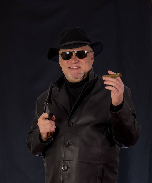 Ένας άντρας με μαύρα ρούχα, μαύρο δερμάτινο παλτό, καπέλο. Γκάνστερ με πούρο και όπλα, ένα πιστόλι του συστήματος Μάουζερ. Μαφιόζι με μαύρα γυαλιά σε σκούρο φόντο. επικίνδυνος, εχθρικός, κακός, αγενής άνθρωπος. Καουμπόης εγκληματίας  - Φωτογραφία, εικόνα