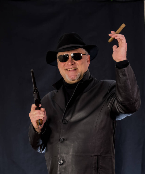 A man in black clothes, a black leather coat, hat. Ganster with a cigar and weapons, a Mauser system pistol. Mafiosi in black glasses on a dark background.  dangerous, unfriendly, evil, rude man. Cowboy felon  - Photo, Image