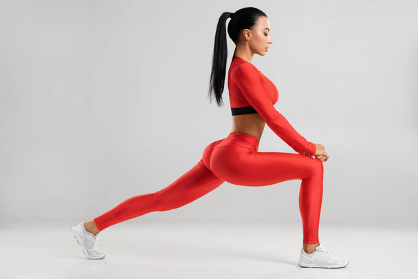 Fitness woman doing lunges exercises for leg muscle workout training. Active girl doing front forward one leg step lunge exercise, on the gray background - 写真・画像