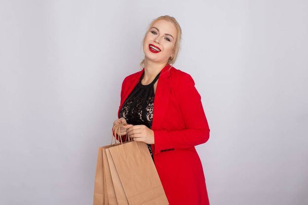 Image of excited blonde woman in red jacket holding brown shopping bags isolated on white bakcground - Fotoğraf, Görsel
