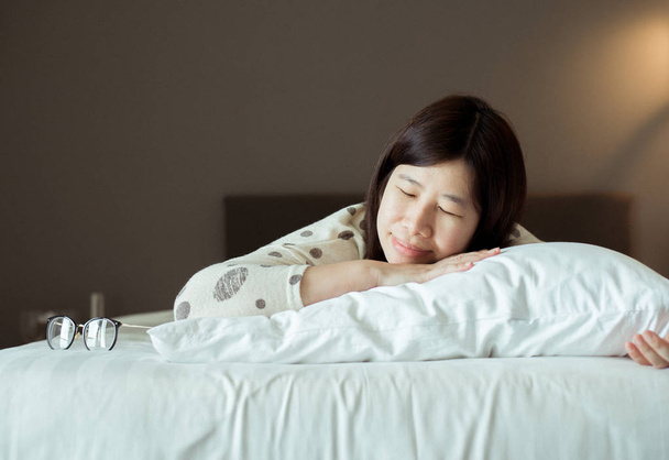 Ασιάτισσα που κοιμάται ευτυχισμένη στο κρεβάτι και χαμογελά με τα χέρια της δίπλα στο πρόσωπό της, γλυκά όνειρα - Φωτογραφία, εικόνα