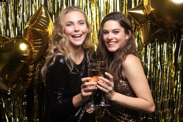 Twee jonge dames die champagne drinken. Afbeelding van meisjes met ballonnen geïsoleerd op gouden achtergrond. - Foto, afbeelding