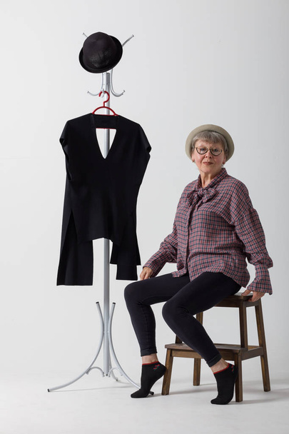 Stylish older woman posing sitting next to hanger on white background. Fashion for older women. - Фото, изображение