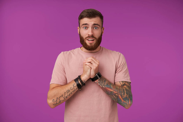 Portrait of pretty blue-eyed bearded brunette guy with tattooes raising folded hands and looking to camera pitifully, wearing beige t-shirt and trendy accessories while posing over purple background - Photo, Image