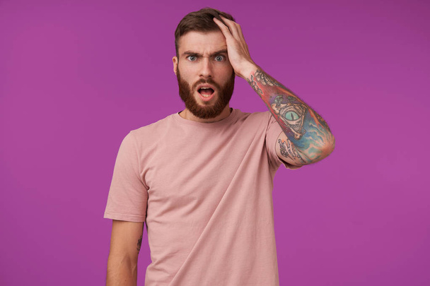 Puzzled pretty young brunette man with beard and tattooes frowning eyebrows with wide mouth opened and keeping raised palm on his head, standing over purple background in beige t-shirt - Photo, Image
