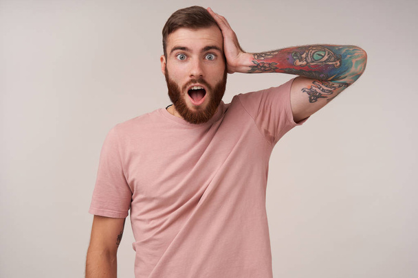 Surprised pretty bearded brunette male with tattooes and trendy haircut clutching his head and looking to camera with wide eyes and mouth opened, isolated over white background - Photo, Image