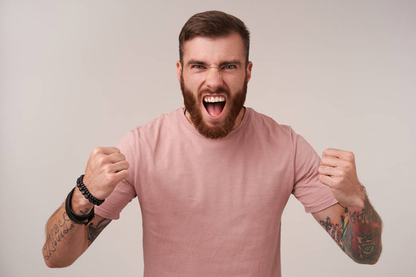 Студийный снимок взволнованного красивого бородатого мужчины с татуировками в бежевой футболке и модных аксессуарах, кричащего громко и складывающего руки кулаками, стоя на белом фоне
 - Фото, изображение