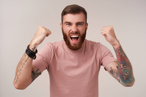 Overjoyed young bearded tattooed male with trendy haircut raising fists happily and screaming with wide mouth opened while watching footbal game, isolated over white background - Foto, immagini