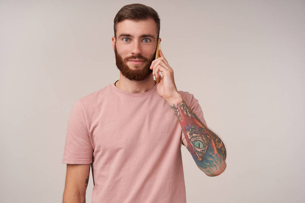 Очаровательный молодой голубоглазый мужчина с татуировками звонит со своего смартфона и смотрит в камеру с позитивным лицом, надевая бежевую футболку, стоя на белом фоне
 - Фото, изображение