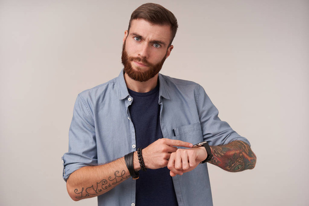 Studio zdjęcie młodego przystojnego brunetka mężczyzna z brodą w niebieskim t-shirt i modne akcesoria, pokazując na zegarku i przypominając o czasie, pozowanie na białym tle - Zdjęcie, obraz