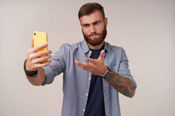 Portré komoly szakállas tetovált srác divatos frizura, amelynek video chat és nézi a kamera súlyos, ráncoló szemöldök csukott ajkak, miközben áll a fehér háttér - Fotó, kép