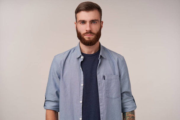 Young handsome bearded blue-eyed brunette with trendy haircut looking at camera with calm face and folded lips, wearing blue t-shirt and shirt while standing over white background - Фото, изображение