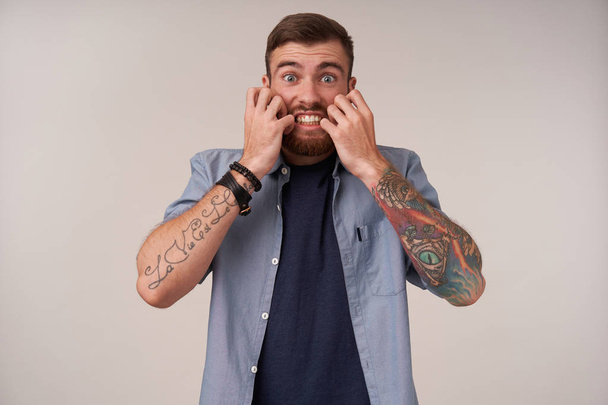 Испуганный молодой татуированный небритый мужчина с короткой стрижкой, держа руки на лице и показывая свои идеальные белые зубы, округлые испуганно глаза, позируя на белом фоне
 - Фото, изображение