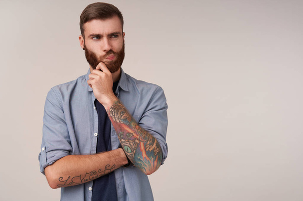 Severe young brunette unshaved male with tattooes holding his chin with raised hand and looking aside, frowning face and twisting his mouth while standing over white background - Foto, Bild