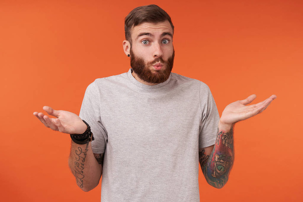 Confused young tattooed man with lush beard shrugging with raised hands, raising eyebrows and pursing lips whille looking at camera, standing over orange background in casual clothes - Photo, Image