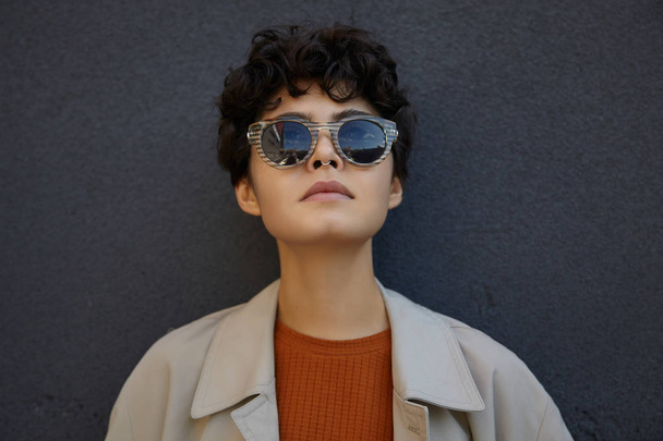 Close-up of attractive dark haired young curly female with short haircut wearing trendy sunglasses, posing outdoor over black concrete wall and looking on sky with folded lips - Photo, Image