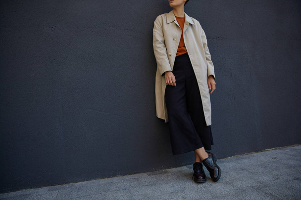 Vystřižená fotka mladé hipsterky oblečené v černých kalhotách, lišáckém svetru a béžovém zákopu, pózující přes černou městskou zeď v módních šatech a kožených brožurách - Fotografie, Obrázek