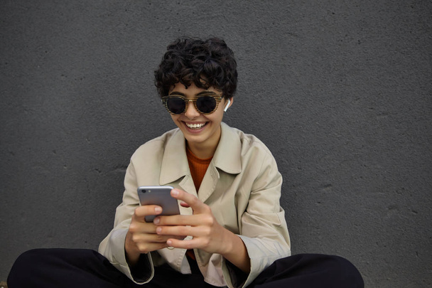 Cheerful pretty curly dark haired female in sunglasses wearing trendy outfit while sitting on city floor in stylish clothes, smiling joyfully while chatting with friends on her smartphone - Foto, Imagem