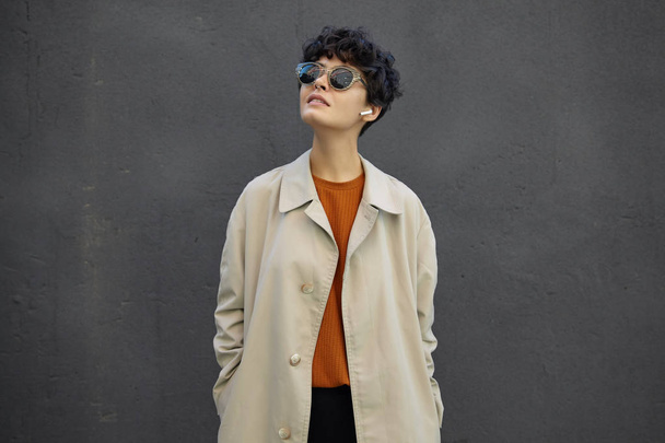 Image of attractive young brunette woman with short curly hair looking upwards with calm face, wearing trendy outfit and sunglasses, standing over city environment - Photo, Image