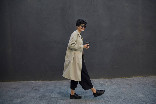 Stylish dark haired hipster female with short curly hair walking in city against black urban wall, wearing black culottes and beige trench coat, holding paper cup with takeaway coffee - Foto, immagini
