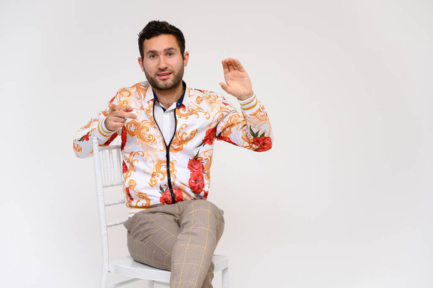 Mens fashion concept. Portrait of a handsome male model, showing hands, wearing a white jacket with a floral pattern, posing on a white background, sidmint on a chair. Black hair. Close Studio Shot - Foto, Bild