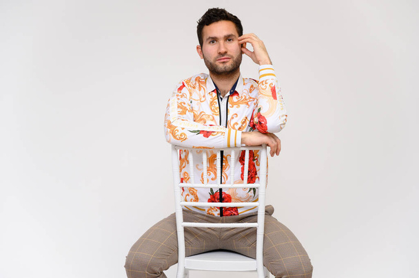 Mens fashion concept. Portrait of a handsome male model, showing hands, wearing a white jacket with a floral pattern, posing on a white background, sidmint on a chair. Black hair. Close Studio Shot - Zdjęcie, obraz