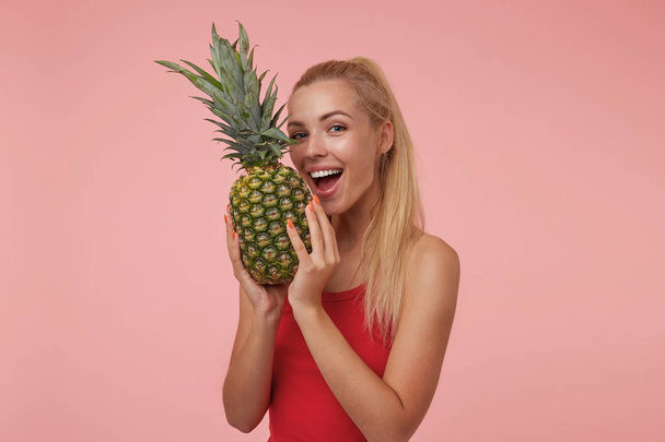 Studio shot of young blonde female with casual hairstyle holding pineapple in her hands, going to bite pineapple, looking at camera and smiling joyfully - Foto, afbeelding