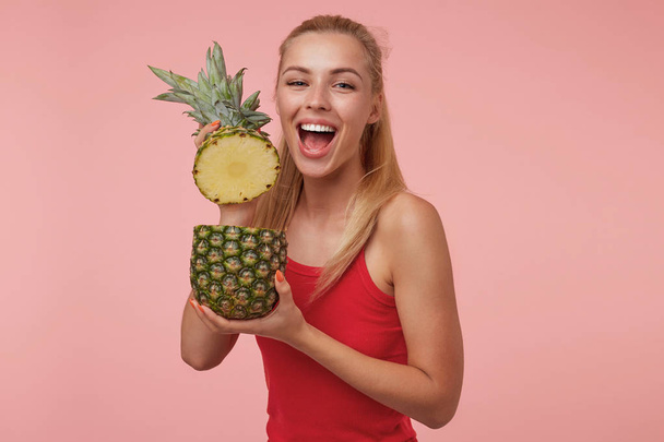 Studio shot of joyful young woman with blonde hair, holding cut pineapple in hands over pink background, looking to camera happily with wide smile - 写真・画像