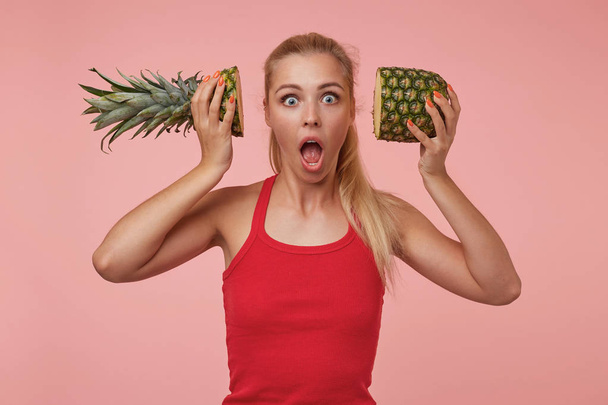 Studio photo of amazed beautiful woman with blonde long hair holding halves of pineapple near her ears, looking at camera with wide eyes and mouth opened - Foto, Bild