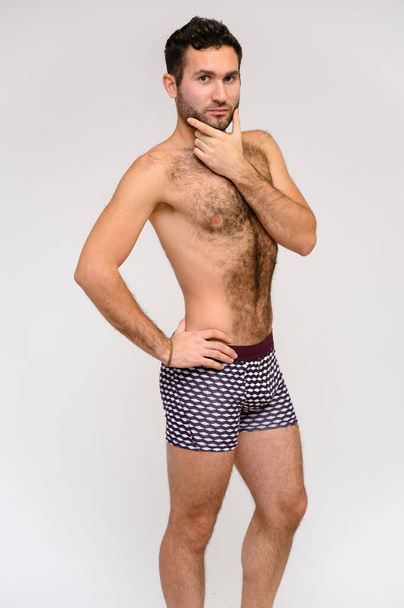 Portrait of a handsome male model with perfect body, standing naked in shorts, posing on a white background. Black hair. Close Studio Shot - Zdjęcie, obraz