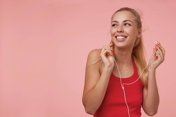 Indoor photo of young pretty lady in red shirt, looking upwards with charming smile, going to listen to music with headphones, posing over pink background - Φωτογραφία, εικόνα