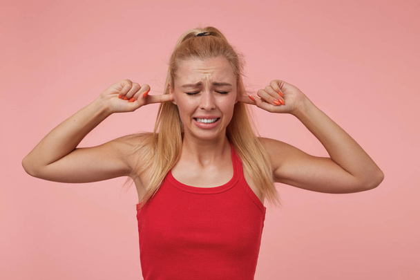 Studiofoto einer gut aussehenden jungen Frau mit lässiger Frisur, die vor rosa Hintergrund steht, die Ohren mit Händen bedeckt und versucht, laute Geräusche zu vermeiden - Foto, Bild