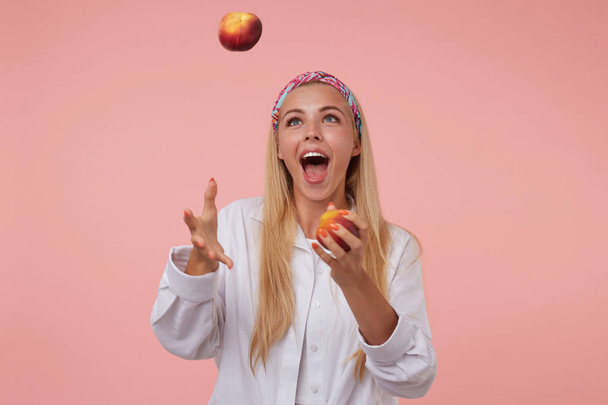Cheerful pretty lady with long blond hair juggling with peaches in studio, having fun over pink background, wearing colored headband and white shirt - Photo, Image
