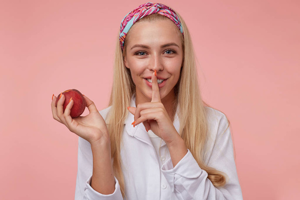 Foto einer jungen hübschen blonden Frau, die lächelt und Schweigegeste zeigt, den Zeigefinger in der Nähe des Mundes hält, zur Ruhe aufruft, vor rosa Hintergrund steht - Foto, Bild