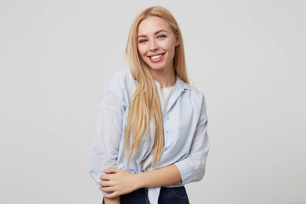 Vnitřní portrét mladé atraktivní blondýny s dlouhými vlasy, usmívající se na kameru upřímně, pózující na bílém pozadí, v džínách a modré košili - Fotografie, Obrázek