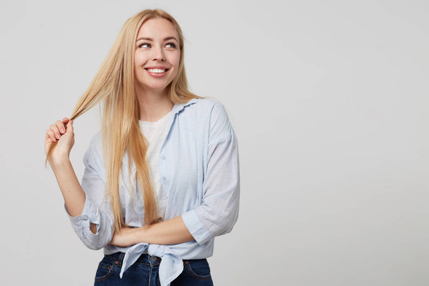 Studio shot of beautiful blonde lady wearing casual clothes, looking aside with broad smile and pulling her hair, isolated over white background - Fotó, kép