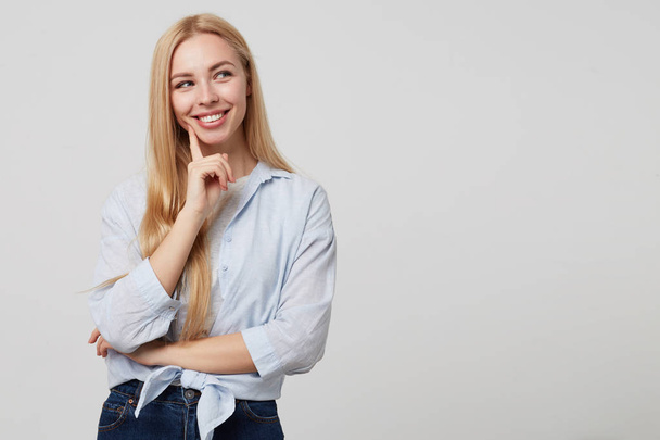 Beautiful young blond lady in casual clothes holding her cheek with index finger and looking joyfully aside, posing over white background - Foto, Imagem