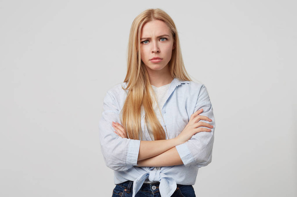 Indoor shot of angry young blonde female with crossed arms wearing casual clothes, looking scowling to camera, posing over white background - Foto, imagen