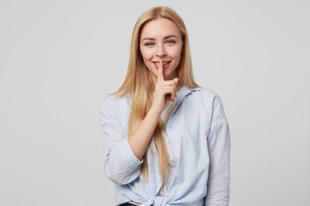 Horizontal shot of beautiful blonde long haired female keeping finger on smiling lips, demonstrating hush sign, asking to keep silence, standing over white background - Fotoğraf, Görsel