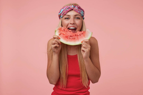 Wewnętrzne ujęcie radosnej, ładnej kobiety z opaską na głowie i długimi blond włosami, pozującej na różowym tle, jedzącej arbuza, będącej w dobrym nastroju - Zdjęcie, obraz